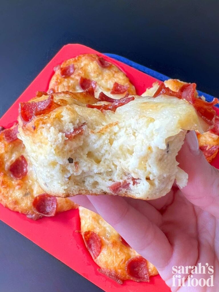 a hand holding a pizza muffin cup with a bite taken out showing the cheesy inside